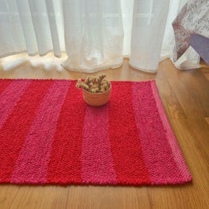 Puede incluir: Una alfombra rectangular con un patrón de rayas en tonos rojos y rosas. Una pequeña cesta tejida llena de flores secas se encuentra sobre la alfombra. La alfombra está colocada sobre un suelo de madera, con cortinas blancas al fondo.