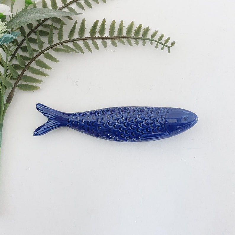 Puede incluir: Una figurita de pescado de cer&aacute;mica azul con un patr&oacute;n de escamas detallado. El pez mide aproximadamente 15 cm de largo y tiene un cuerpo ligeramente curvado.