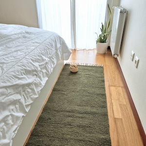 Op de afbeelding: Een geweven groen tapijt met een franje rand, geplaatst op een houten vloer in een slaapkamer. Een wit bed met een wit dekbed is op de achtergrond.