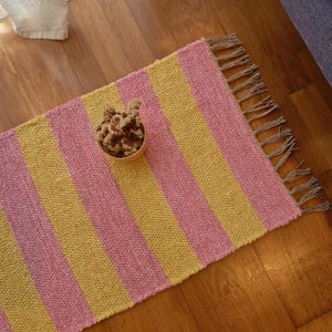 May include: A rectangular rug with vertical stripes in alternating shades of pink and yellow. The rug has a fringed edge and is made of a woven material. A small bowl of dried flowers sits on the rug.