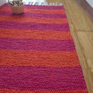 May include: A vibrant, hand-woven rug with alternating horizontal stripes of bright pink and orange. The rug has a textured appearance and a fringed edge. The rug is placed on a wooden floor.