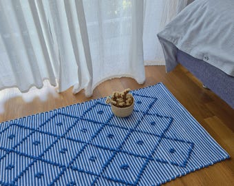 May include: A blue and white striped rug with a geometric diamond pattern. The rug features a textured design with raised blue elements. A small woven basket filled with dried flowers sits on the wooden floor near the rug.