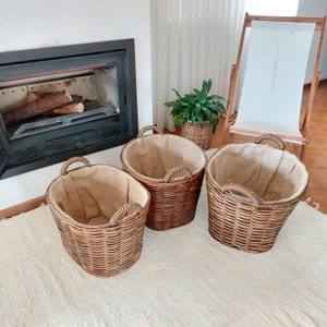 May include: Three brown wicker baskets with woven handles and a burlap lining. The baskets are oval-shaped and vary slightly in size. They are placed on a cream-colored rug, near a fireplace and a wooden chair.