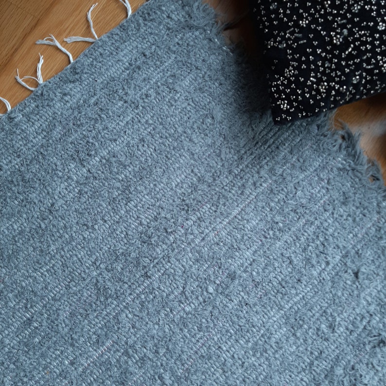 May include: A close-up of a gray, textured rug with a frayed edge. The rug has a subtle striped pattern.