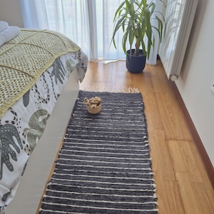 Könnte beinhalten: Ein dunkelgrauer rechteckiger Teppich mit weißen horizontalen Streifen. Der Teppich liegt auf einem Holzboden in der Nähe eines Bettes mit einer gemusterten Bettdecke und einer Topfpflanze. Ein kleiner Weidenkorb mit Trockenblumen steht auf dem Teppich.