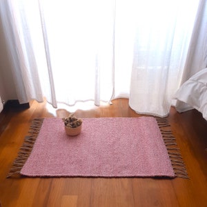 Puede incluir: Una alfombra tejida de color rosa con flecos en los bordes. La alfombra está sobre un suelo de madera y hay un pequeño cuenco con flores secas en el centro de la alfombra.
