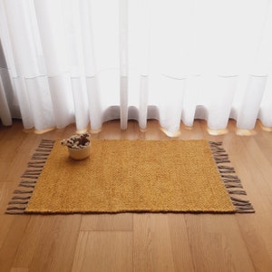 Puede incluir: Una alfombra tejida de color amarillo mostaza con flecos en todos los lados. La alfombra está sobre un suelo de madera delante de una cortina blanca transparente.