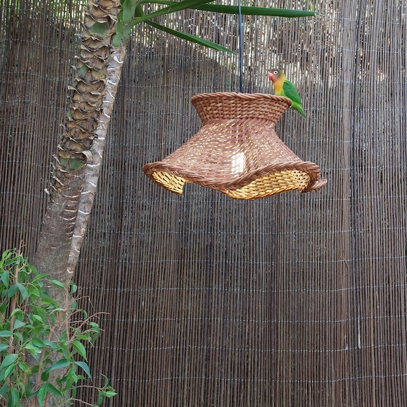 May include: A brown woven pendant lamp with a flared shade, hanging outdoors. A small, colorful bird figurine perches on top. The lamp is suspended in front of a bamboo fence and a palm tree.