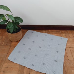 Puede incluir: Una alfombra tejida de color gris claro con un patr&oacute;n texturizado de peque&ntilde;os c&iacute;rculos. La alfombra est&aacute; sobre un suelo de madera.