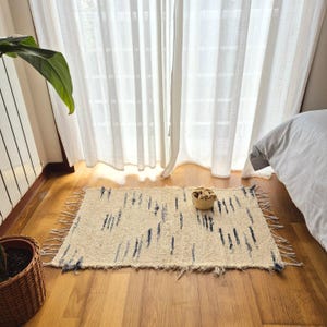 Puede incluir: Una alfombra rectangular blanquecina con flecos y un patrón de rayas verticales azul oscuro. Una pequeña cesta tejida está sobre la alfombra. La alfombra está sobre un suelo de madera cerca de una ventana con cortinas blancas.