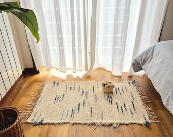 May include: An off-white rectangular rug with a fringe and a pattern of dark blue vertical lines. A small woven basket is on the rug. The rug is on a wooden floor near a window with white curtains.
