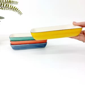 Pode incluir: Quatro pratos retangulares de cer&acirc;mica nas cores amarelo, turquesa, laranja e azul. Cada prato tem um interior branco e uma borda preta. Um prato amarelo &eacute; levantado, mostrando o design.