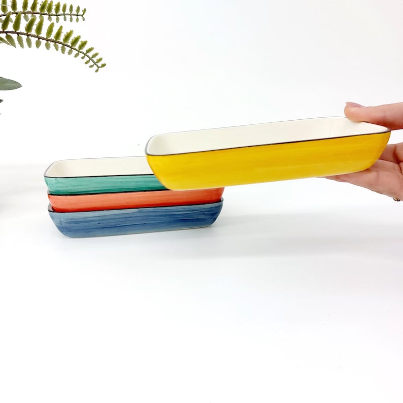 Pode incluir: Quatro pratos retangulares de cer&acirc;mica nas cores amarelo, turquesa, laranja e azul. Cada prato tem um interior branco e uma borda preta. Um prato amarelo &eacute; levantado, mostrando o design.