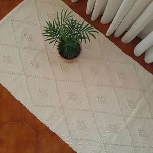 May include: A white woven rug with a diamond pattern and a small potted plant in the center.