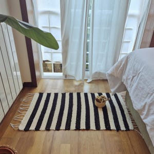 Puede incluir: Una alfombra rectangular que presenta un patrón repetitivo de gruesas rayas verticales en color negro y blanco roto. La alfombra tiene una superficie tejida con textura y está rematada con largos flecos blancos sueltos en los extremos cortos.
