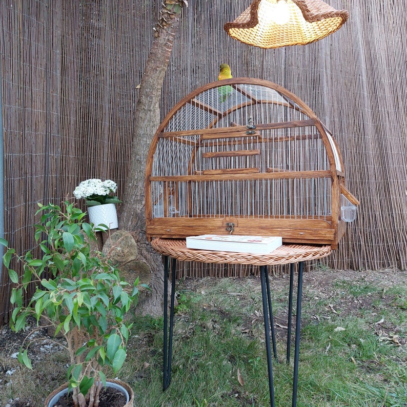 May include: A wooden birdcage with a brown finish and a wire mesh top. The cage is sitting on a small table with a wicker top and black metal legs. A small bird is perched on the top of the cage.
