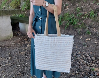 May include: A white tote bag with fringe trim and a striped pattern. The bag is made of a woven fabric and has a brown leather strap.