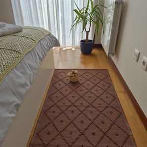 Könnte beinhalten: Ein brauner rechteckiger Teppich mit einem Rautenmuster und kleinen, erhabenen Punkten. Der Teppich liegt auf einem Holzboden. Ein kleiner Weidenkorb mit dekorativen Gegenständen steht auf dem Teppich. Ein Bett mit einer grünen Decke ist im Hintergrund.