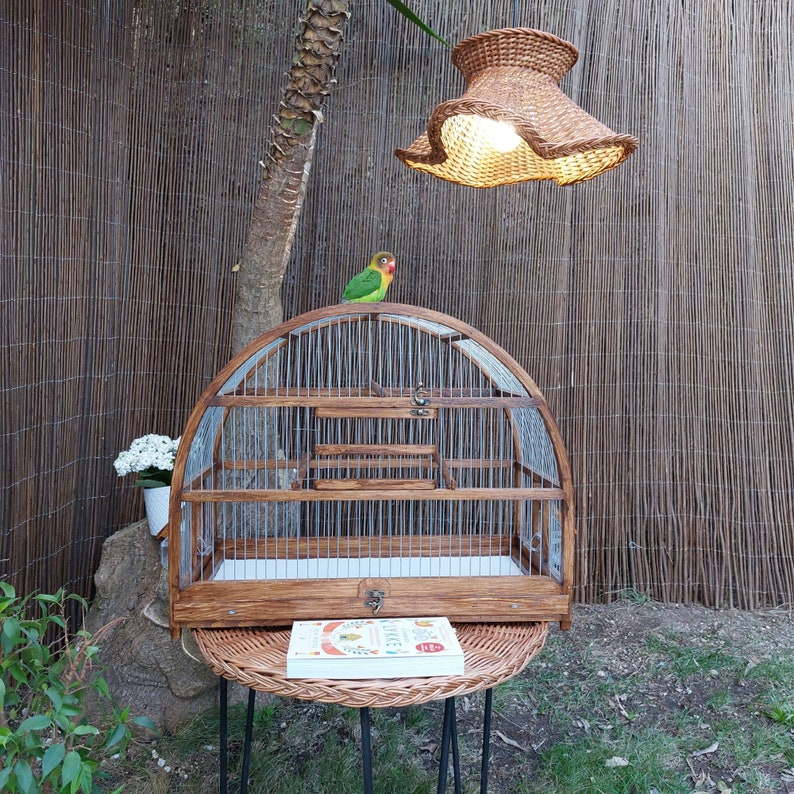 May include: A wooden birdcage with a metal wire top and a small green and red bird perched on the top. The cage is sitting on a wicker table with a book on top. A woven lampshade hangs above the cage.