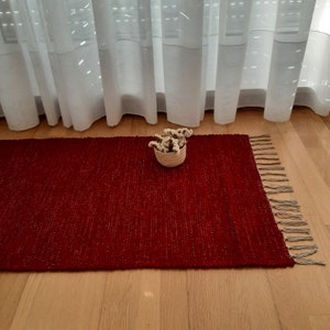 Peut inclure: Un tapis tissé rouge avec des franges sur les bords. Le tapis est placé sur un sol en bois et a un petit panier de fleurs blanches au centre.