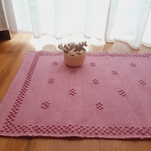 Pu&ograve; includere: Un tappeto rosa tessuto con un motivo floreale e un bordo a perline. Un piccolo cestino intrecciato pieno di fiori secchi &egrave; posto sul tappeto. Il tappeto &egrave; su un pavimento in legno vicino a una finestra con tende trasparenti.