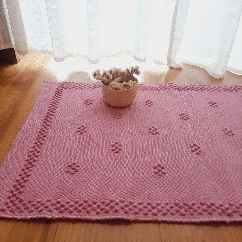 Pu&ograve; includere: Un tappeto rosa tessuto con un motivo floreale e un bordo a perline. Un piccolo cestino intrecciato pieno di fiori secchi &egrave; posto sul tappeto. Il tappeto &egrave; su un pavimento in legno vicino a una finestra con tende trasparenti.