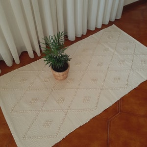 May include: A white woven rug with a diamond pattern and raised pom-poms. The rug is on a brown tiled floor and a potted plant is in the foreground.