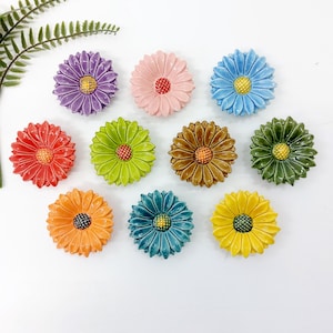 Puede incluir: Una colección de decoraciones de flores de margarita de cerámica en varios colores, incluyendo morado, rosa, azul, rojo, verde, marrón, naranja, turquesa y amarillo. Cada flor tiene un centro contrastante.
