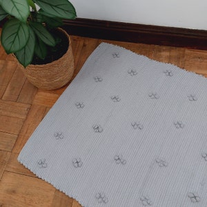 Puede incluir: Una alfombra gris claro con una superficie texturizada y un patr&oacute;n repetitivo de peque&ntilde;as flores en relieve. La alfombra est&aacute; hecha de un material suave y tejido.