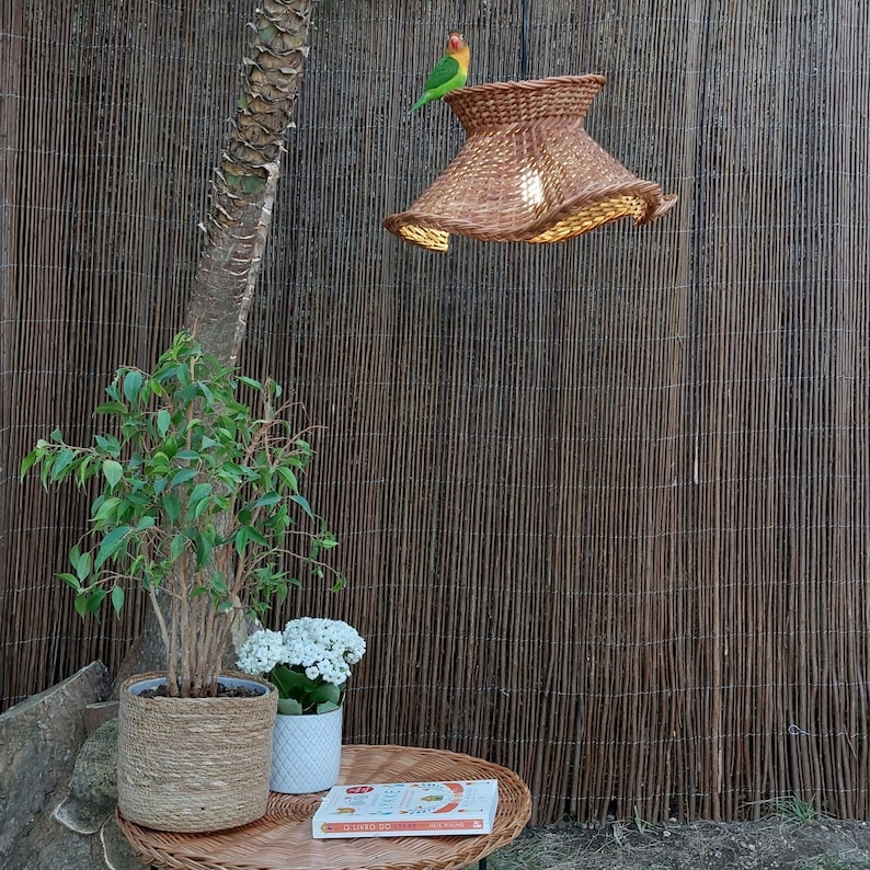 May include: A woven brown lampshade with a flared design hangs from the ceiling. A small, colorful bird perches on top. The lampshade is lit from within, casting a warm glow. A small table with a plant and a book is in the foreground.