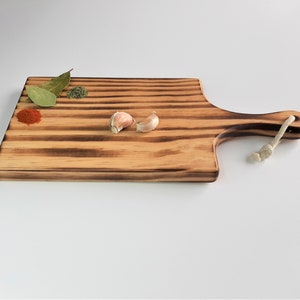 May include: A rectangular wooden chopping board with a handle and a rope loop. The board has a dark brown wood grain and is striped with lighter wood. There are two cloves of garlic, a small pile of red pepper flakes, and a few sprigs of bay leaves on the board.