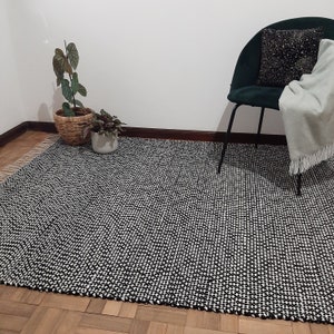 May include: Black and white patterned rug with a woven texture. The rug is laid out on a wooden floor with a green chair and plants in the background.