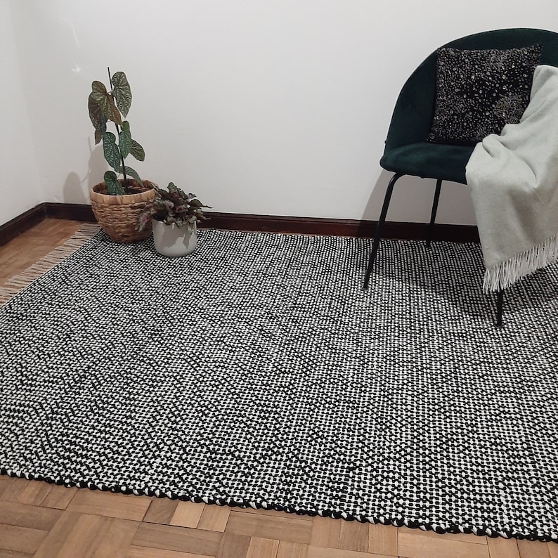 May include: Black and white patterned rug with a woven texture. The rug is laid out on a wooden floor with a green chair and plants in the background.