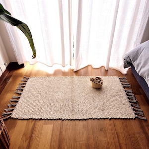 May include: A rectangular, beige woven rug with a textured surface and fringed edges. The rug is placed on a wooden floor near a window with white curtains. A small woven basket with dried flowers sits on the rug.