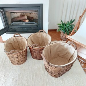 May include: Three woven wicker baskets with rope handles. Each basket has a burlap lining. The baskets are different sizes and are arranged on a white rug. The background includes a fireplace with logs and a plant.