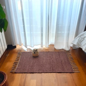 May include: A brown woven rug with fringe edges. The rug is laid out on a wooden floor in front of a white sheer curtain.