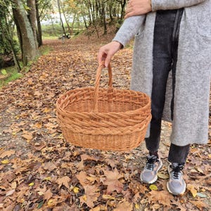 May include: A woven wicker basket with a handle, in a natural brown color. The basket is oval shaped and has a layered design. The basket is sitting on a bed of fallen autumn leaves.