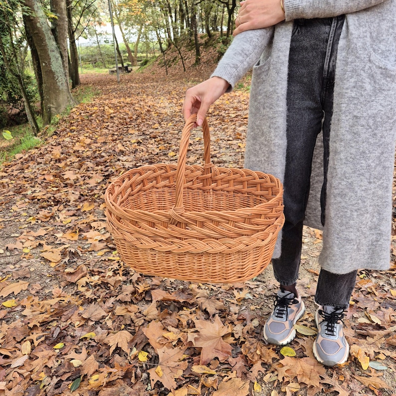 May include: A woven wicker basket with a handle, in a natural brown color. The basket is oval shaped and has a layered design. The basket is sitting on a bed of fallen autumn leaves.