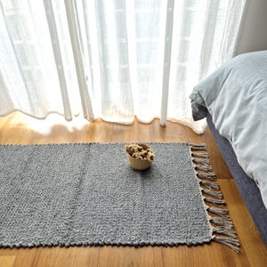 Puede incluir: Una alfombra rectangular gris con una superficie texturizada y bordes con flecos. Una pequeña cesta tejida llena de flores secas está sobre la alfombra. La alfombra está colocada sobre un suelo de madera cerca de una cama y una ventana con cortinas transparentes.