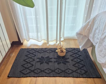 Puede incluir: Una alfombra rectangular tejida en color negro que presenta un patrón geométrico en relieve de rombos y dos motivos centrales en forma de estrella. La alfombra tiene una superficie texturizada de color oscuro uniforme y sirve de base.