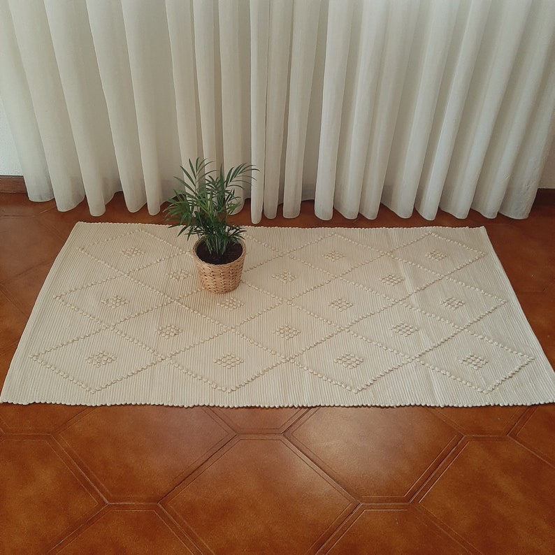 May include: A white woven rug with a diamond pattern. The rug is on a brown tiled floor and a potted plant is in the center of the rug.