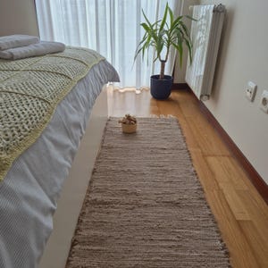 May include: A rectangular, woven, beige rug with a fringed edge is placed on a hardwood floor. The rug is positioned near a bed with a white base and a light green and white patterned blanket. A small basket sits on the rug.