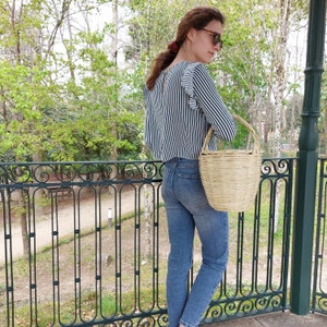 K&ouml;nnte beinhalten: Eine Frau tr&auml;gt ein gestreiftes schwarz-wei&szlig;es Oberteil und eine blaue Jeans und tr&auml;gt eine geflochtene Korbtasche.