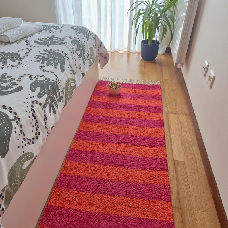 May include: A vibrant, striped rug in shades of pink and orange. The rug is rectangular and placed on a wooden floor, with a bed and a plant in the background. The rug's design features alternating bands of bright pink and orange.
