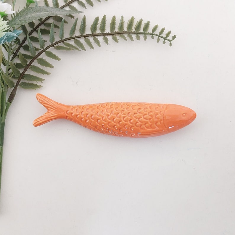 Puede incluir: Una figurita de pescado de cer&aacute;mica naranja con un patr&oacute;n de escamas detallado. El pez mide aproximadamente 13 cm de largo y tiene un cuerpo ligeramente curvado.