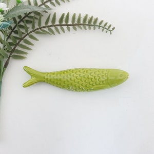 Puede incluir: Una figurita de pescado de cer&aacute;mica verde con una superficie texturizada. El pez tiene un cuerpo ligeramente curvado y una cola puntiaguda.