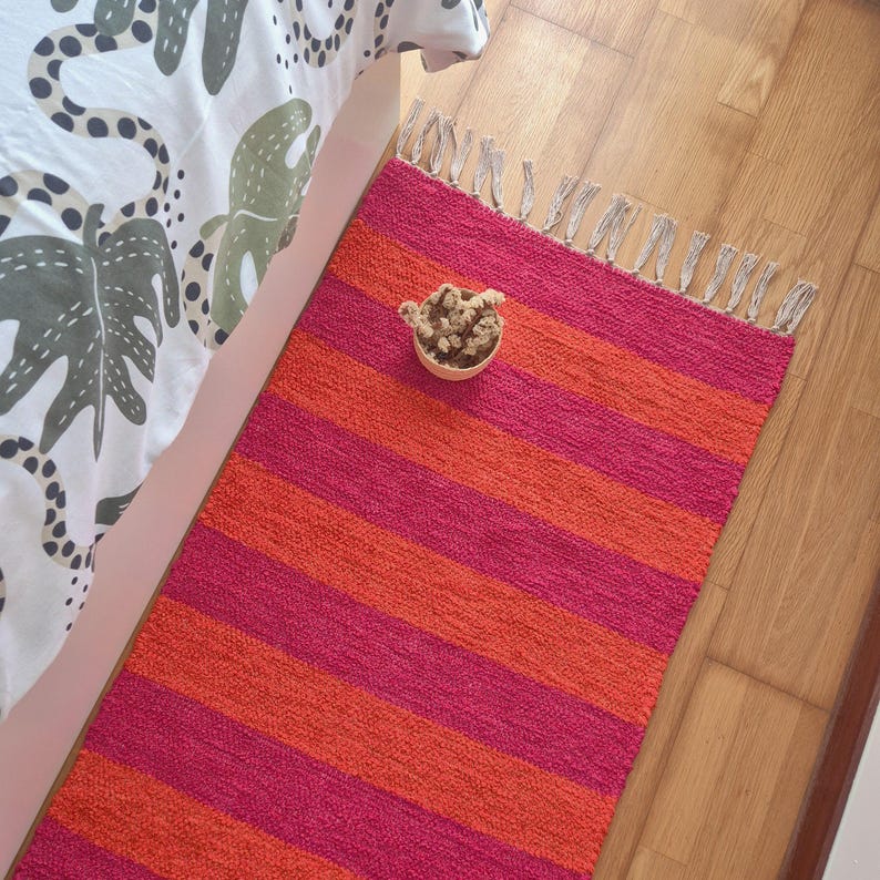 May include: A rectangular rug with alternating horizontal stripes of bright pink and orange. The rug has a fringed edge and is placed on a wooden floor. A small, round, beige bowl with dried flowers sits on the rug.