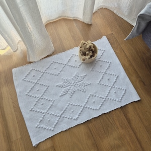 Può includere: Un tappeto rettangolare bianco con un motivo geometrico a rombi e stelle. Il tappeto è realizzato in un materiale strutturato ed è posizionato su un pavimento in legno. Un piccolo cestino intrecciato con fiori secchi è appoggiato sul tappeto.