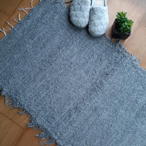 May include: A gray, fuzzy rug with a frayed edge. Two gray slippers are on top of the rug.