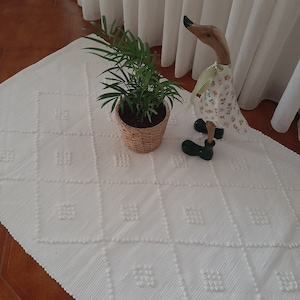 Puede incluir: Una alfombra blanca texturizada con un patrón de diamante. Una planta en maceta y una figura de pato de madera están sobre la alfombra.